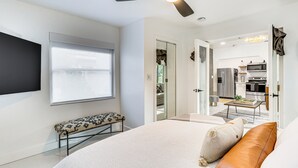 Ein modernes Schlafzimmer mit einem großen Bett, einer Bank, einem Deckenventilator und Blick zur Küche durch eine offene Tür.