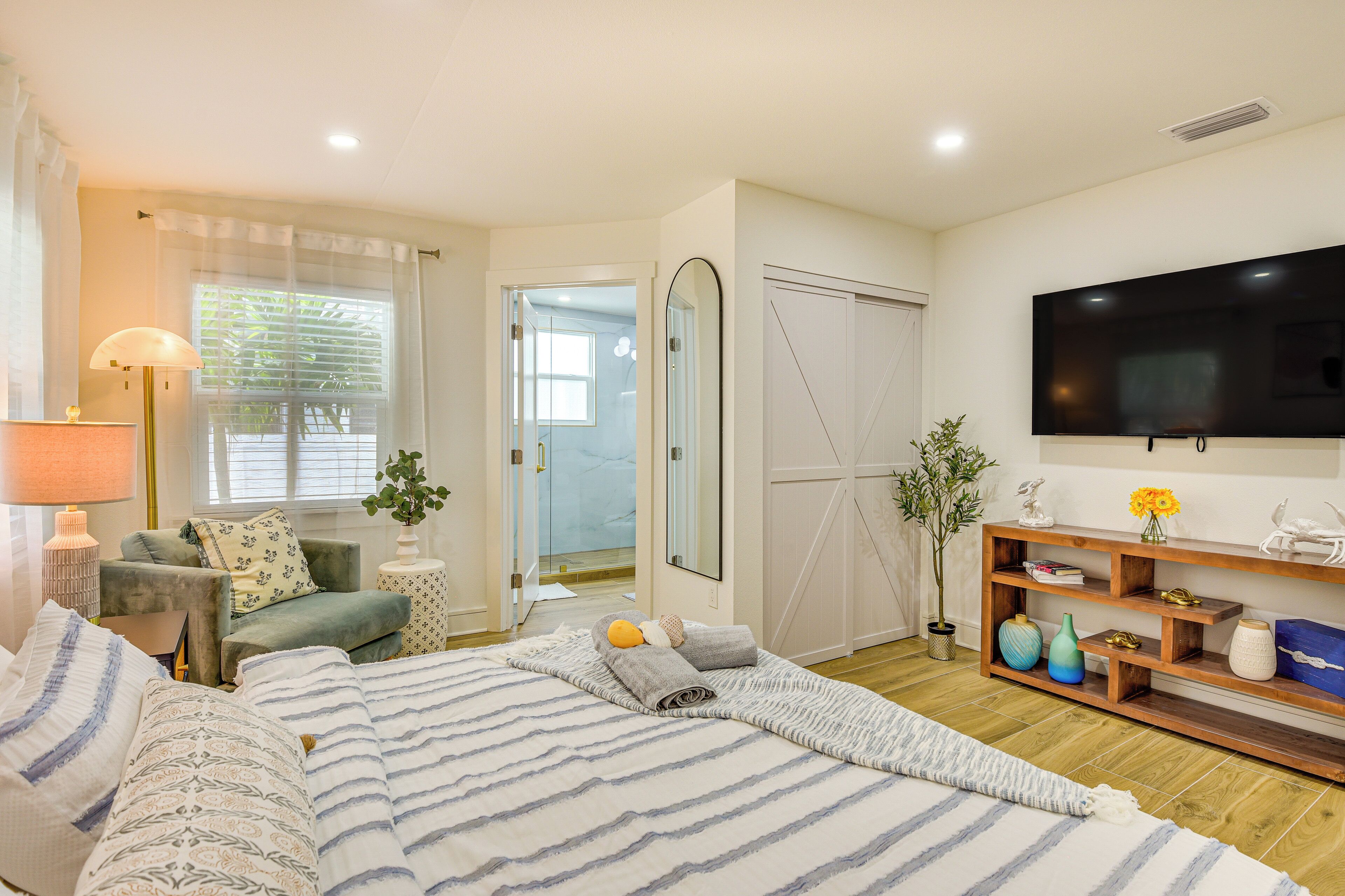 A bedroom with a large bed, a TV, a sofa, a mirror, and a wooden shelf with decorative items.