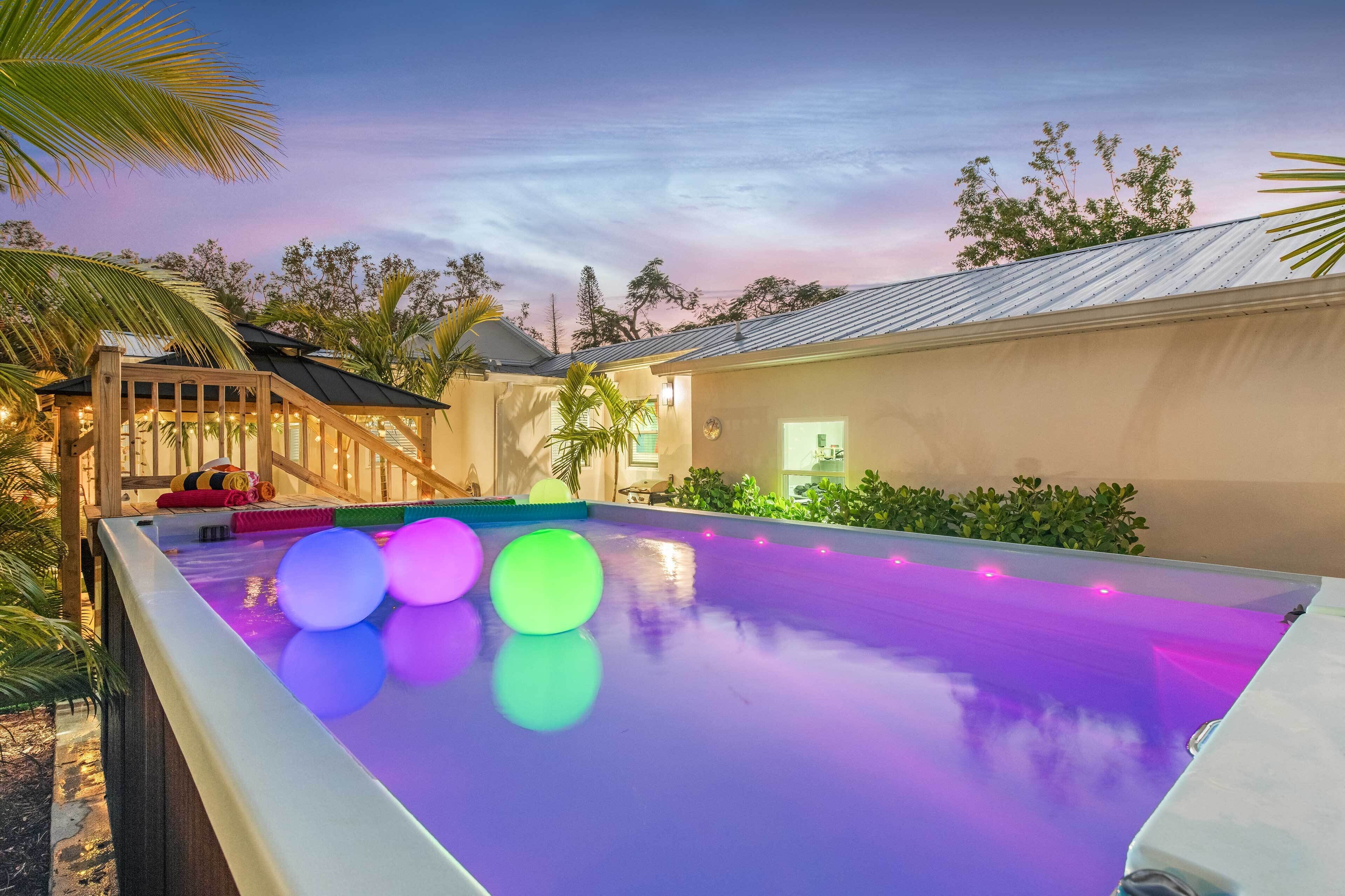 A swimming pool with colorful lights, surrounded by greenery and a building with a sloped roof.