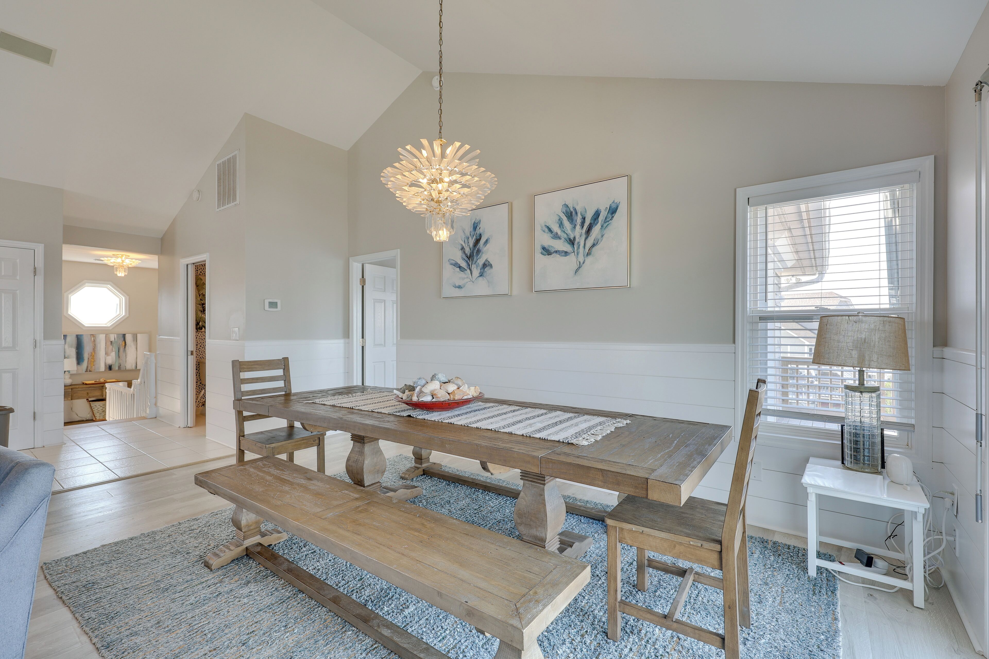 A dining area with a wooden table, benches, and a chandelier. There are framed artworks on the wall and a window with blinds.