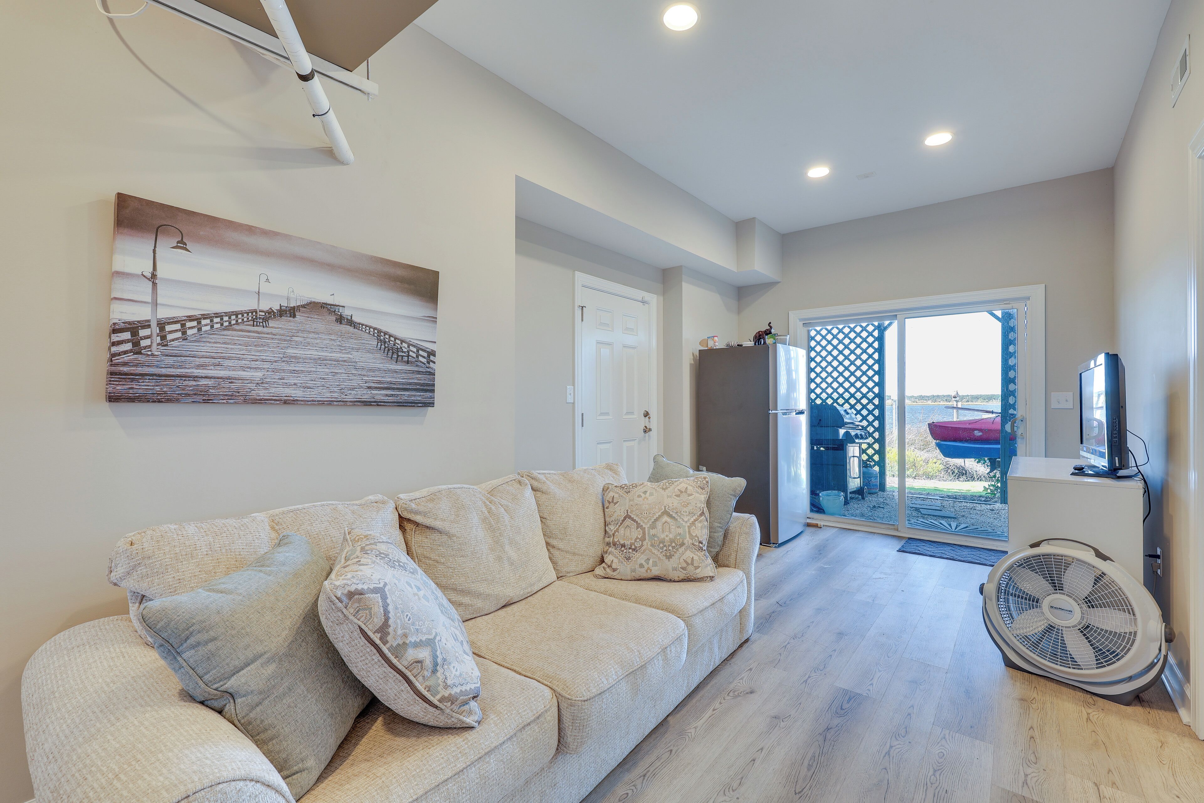A living room with a beige sofa, a painting of a pier, and a fan.
