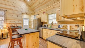 A wooden cabin interior with a kitchen area featuring wooden cabinets, a refrigerator, and a dining table with chairs. - Near Greenbrier River! Mtn-view Cabin w/ Fire Pit (Marlinton)