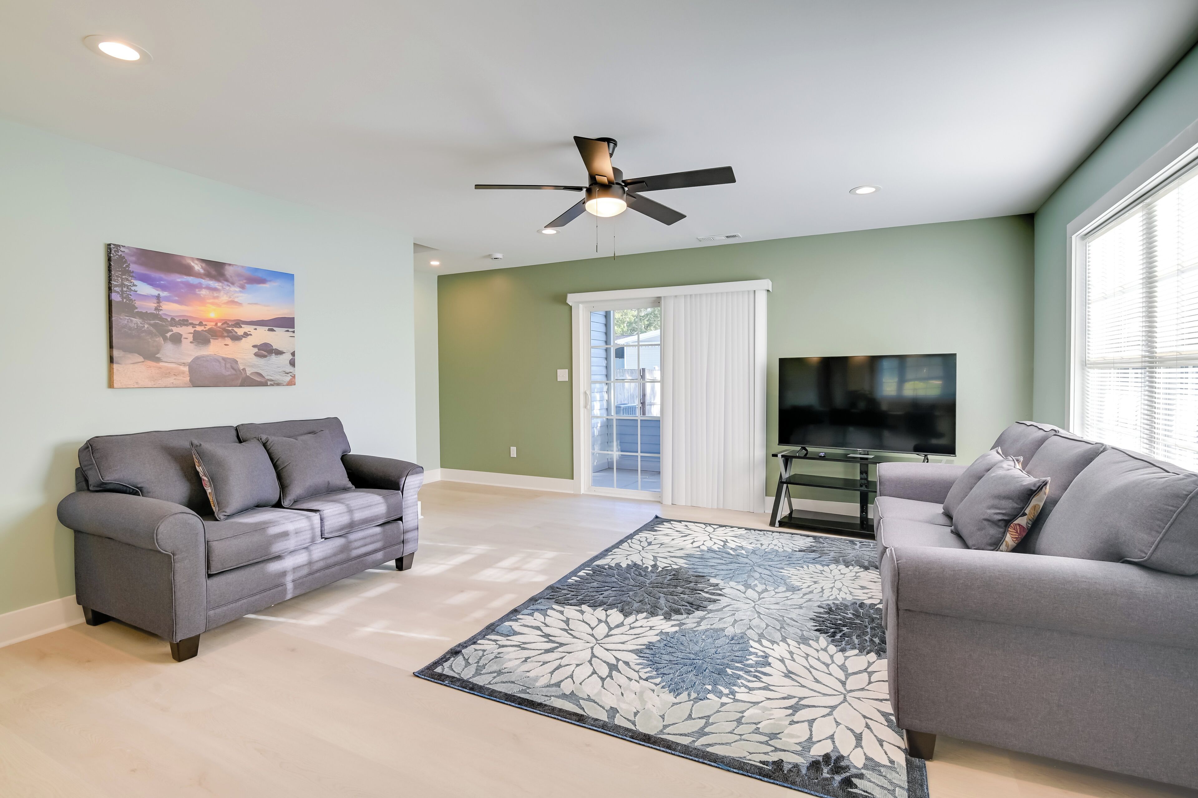Un salon avec deux canapés gris, un grand tapis à motifs floraux, un ventilateur de plafond, un tableau représentant un coucher de soleil et un téléviseur fixé au mur.