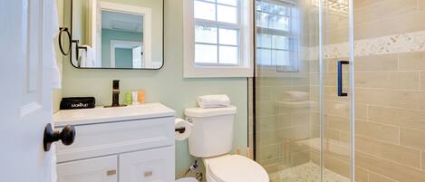 A bathroom with a toilet, a sink with a makeup bag, a trash bin, a shower with a glass door, and a window with blinds.