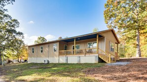 A single-story wooden cabin with a covered porch and a small deck, surrounded by trees and a dirt path. - Lake-view Home in Statham w/ On-site Fishing (Statham)