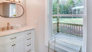 A bathroom with a marble countertop, a large oval mirror, a sink with a single faucet, and a window with a view of a wooden deck and greenery. - Lake-view Home in Statham w/ On-site Fishing (Statham)