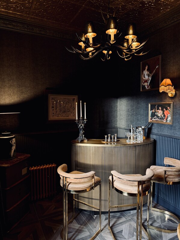 A bar area with a round bar, three bar stools, a chandelier, and wall-mounted artwork. - Lodges On Loch Ness (Inverness)