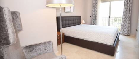 A bedroom with a bed, a bedside table, a lamp, a chair, and a window with curtains.
