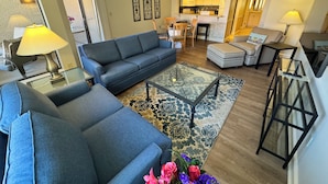 A modern living room with a sofa, coffee table, and lamps. There is a kitchen area with cabinets and a dining table.