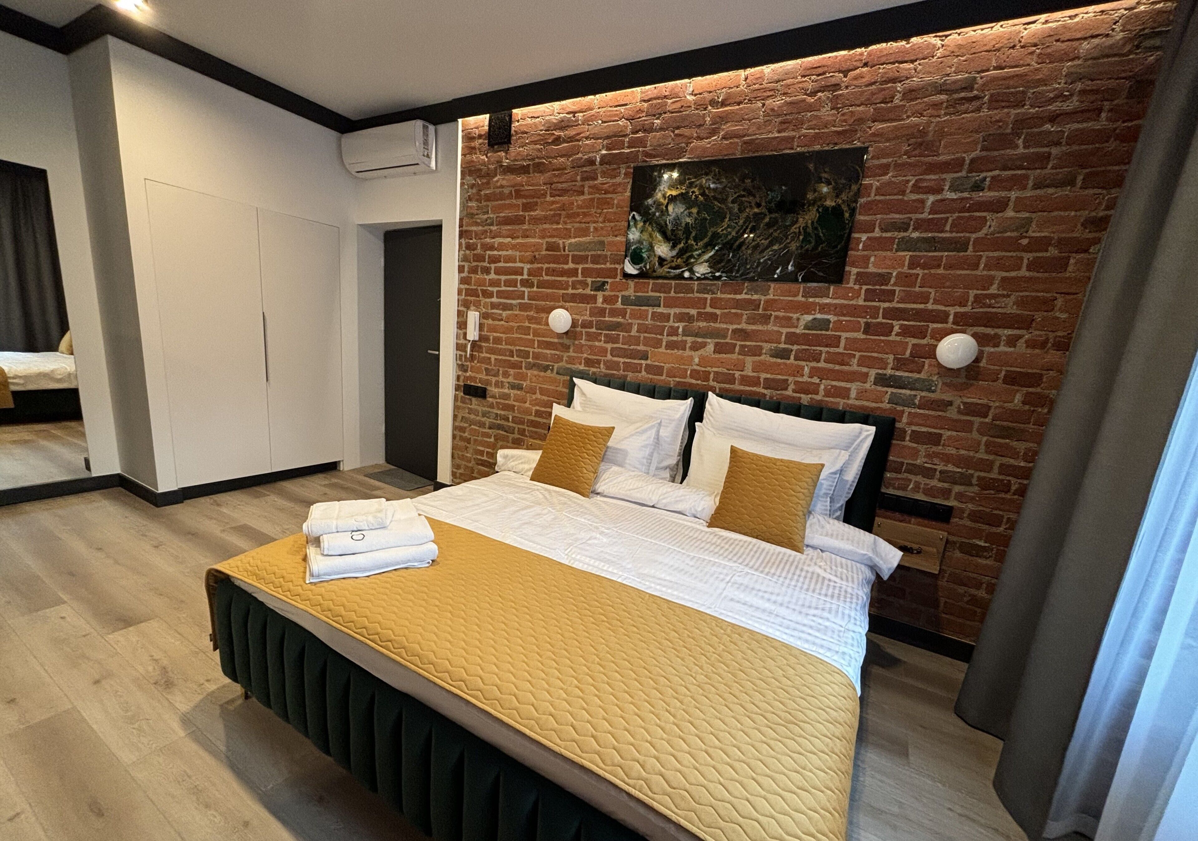 A modern hotel room with a brick wall, a large bed with white bedding and a mustard-colored quilt, a headboard, and a painting above the bed.