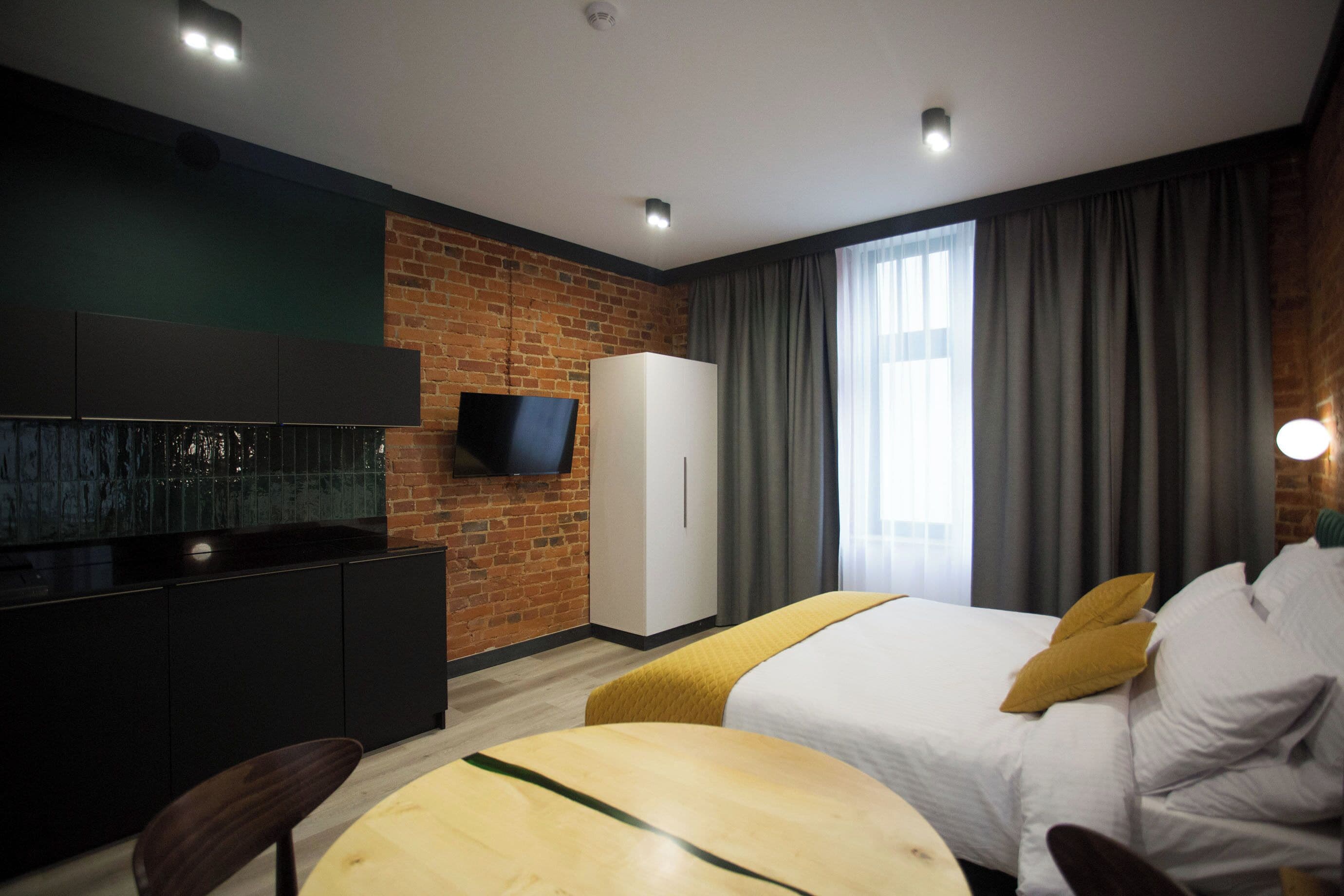 A modern hotel room with a brick wall, a flat-screen TV, a bed with white bedding and yellow pillows, a small round table, and a wardrobe.