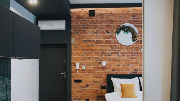 A modern hotel room with a brick wall, a bed with a yellow throw, a round mirror, and a dark headboard.