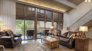 A spacious living room with a large window, a wooden coffee table, leather sofas, and a staircase. - High Mountain Home | AC, Pool, Hot Tub, Golf & Ski (Durango)