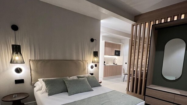 A modern bedroom with a large bed, bedside tables, a built-in TV, and a small kitchenette. - INTRAMUROS Quiet Apartments (Muros)