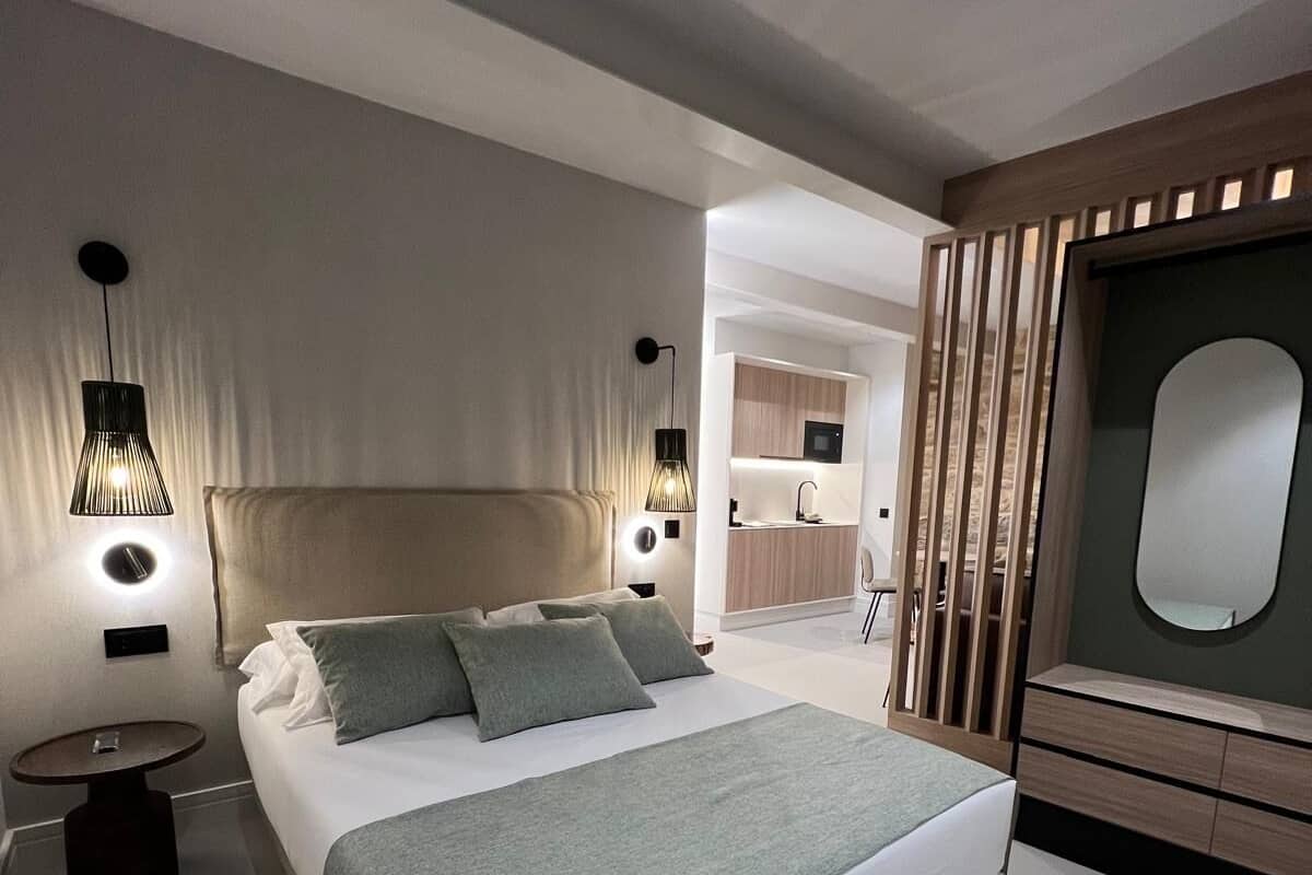 A modern bedroom with a large bed, bedside tables, a built-in TV, and a small kitchenette.