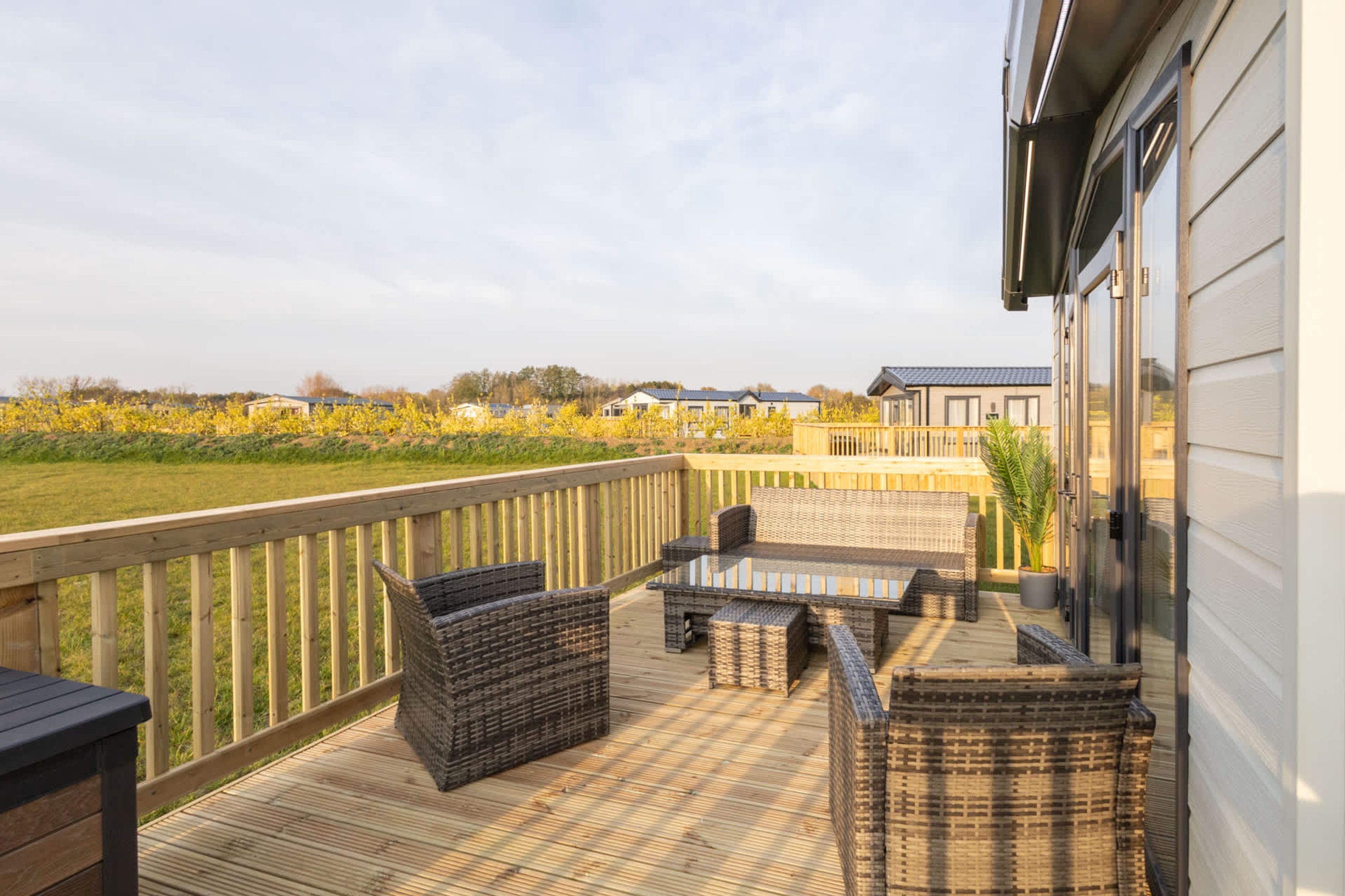 Una terraza de madera con muebles de mimbre, una mesa de vidrio y vista a un campo y edificios a lo lejos.