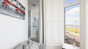A room with a grey sofa, a small table with a bottle and glass, a painting of red umbrellas, and a view of a road and buildings through a window. - Host Stay Egton Bridge at the Sandbeck (Whitby)