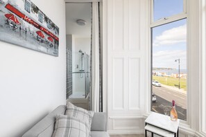 A room with a grey sofa, a small table with a bottle and glass, a painting of red umbrellas, and a view of a road and buildings through a window. - Host Stay Egton Bridge at the Sandbeck (Whitby)