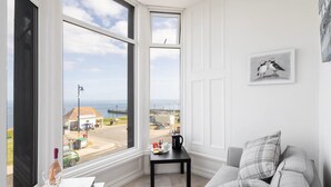 A room with a large window offering a view of the sea and a road. There's a white table with a bottle and glasses, a black table with a kettle, and a grey sofa with pillows. - Host Stay Egton Bridge at the Sandbeck (Whitby)