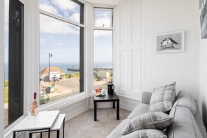 A room with a large window offering a view of the sea and a road. There's a white table with a bottle and glasses, a black table with a kettle, and a grey sofa with pillows. - Host Stay Egton Bridge at the Sandbeck (Whitby)