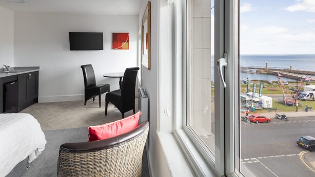 Et hotelværelse med seng, spisebord med stole, fjernsyn ophængt på væggen og udsigt til havet og en mole.