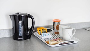 En køkkenbordplade med en sort kaffemaskine, en hvid kop, en bakke med mad og en dåse.
