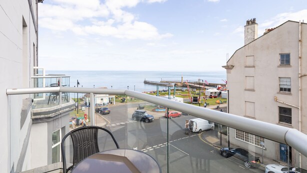 A balcony with a table and chairs overlooking a street with parked cars and a view of the sea. - Host Stay Grosmont at the Sandbeck (Whitby)
