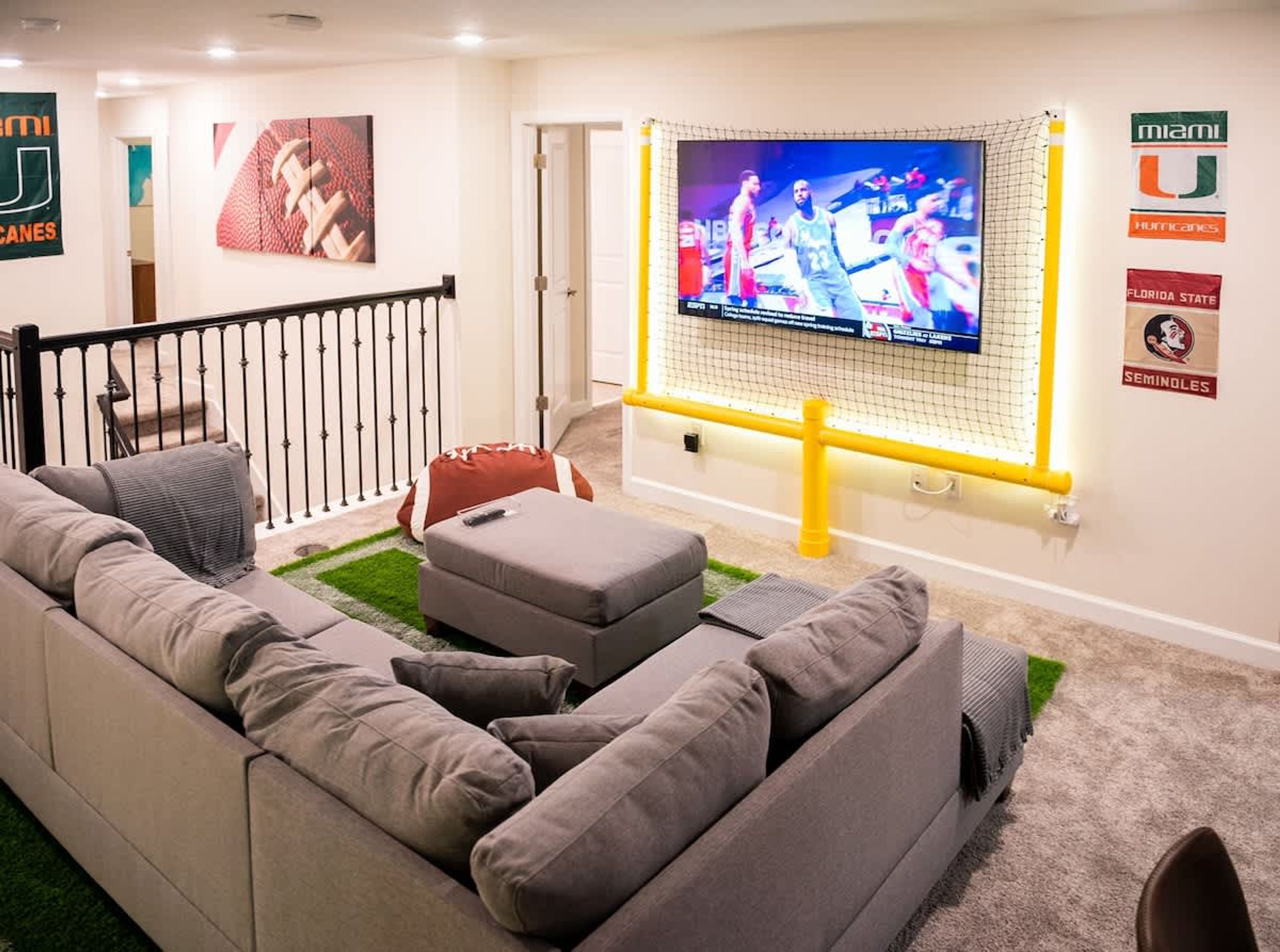 A living room with a large flat-screen TV mounted on a yellow stand, a grey sectional sofa, and a small ottoman.