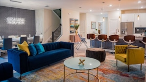Ein modernes Wohnzimmer mit einem blauen Sofa, einem gelben Sessel und einem Glastisch. Eine Treppe führt zu einer Küchenzeile mit weißen Schränken und einem Edelstahlkühlschrank.