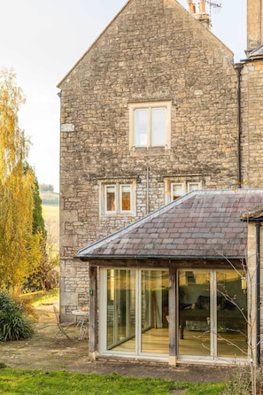 A stone building with a sloped roof, large glass doors, and a small patio area with outdoor furniture. - Georgian House With Garden Views Tennis Court (Bath)