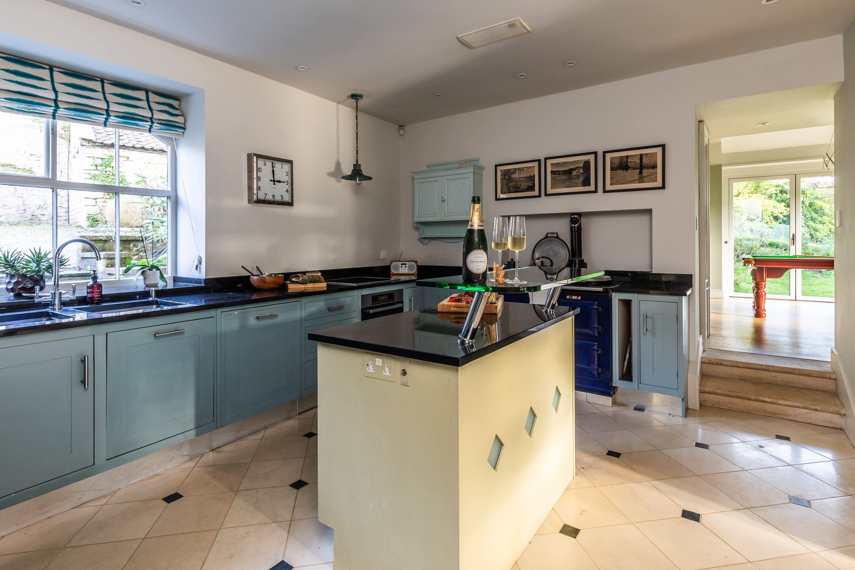 A modern kitchen with light blue cabinets, a central island, and a view of the garden through large windows.