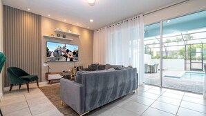 A modern living room with a grey sofa, a TV mounted on the wall, a green chair, and a view of a swimming pool through large windows. - 111 Amazing Townhouse For Your Family a Relaxing Vacation (Davenport)