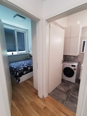 Ein kleiner Raum mit einem Bett, einer Waschmaschine und einem kleinen Fenster mit Jalousien.