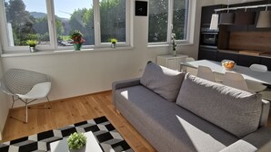 A modern living room with a grey sofa, a white coffee table, and a black-and-white patterned rug. Large windows offer a view of greenery and buildings. - Apartments Egis Koper (Koper)
