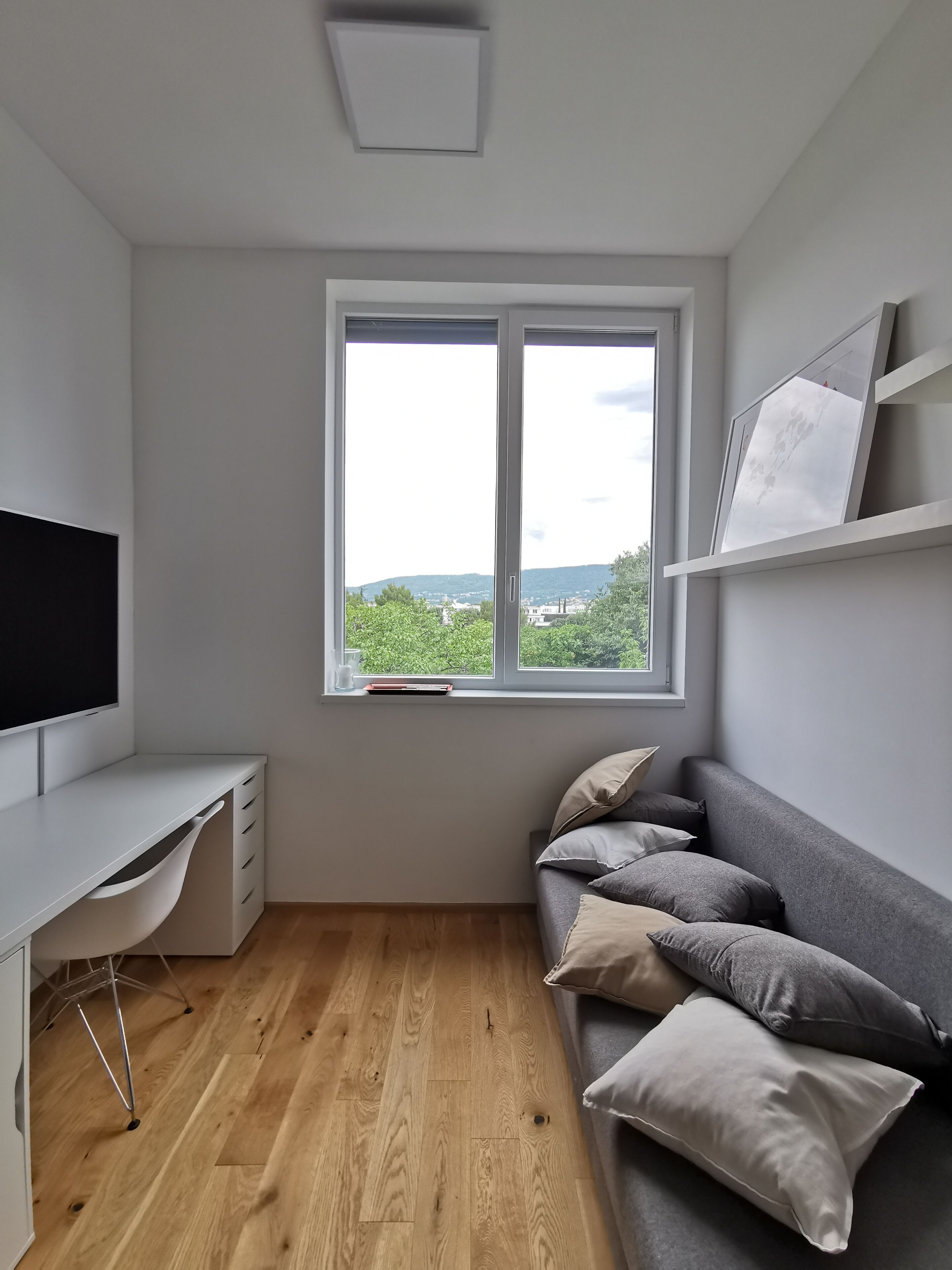 A modern room with a white desk, a grey sofa, and a window offering a view of greenery and buildings.