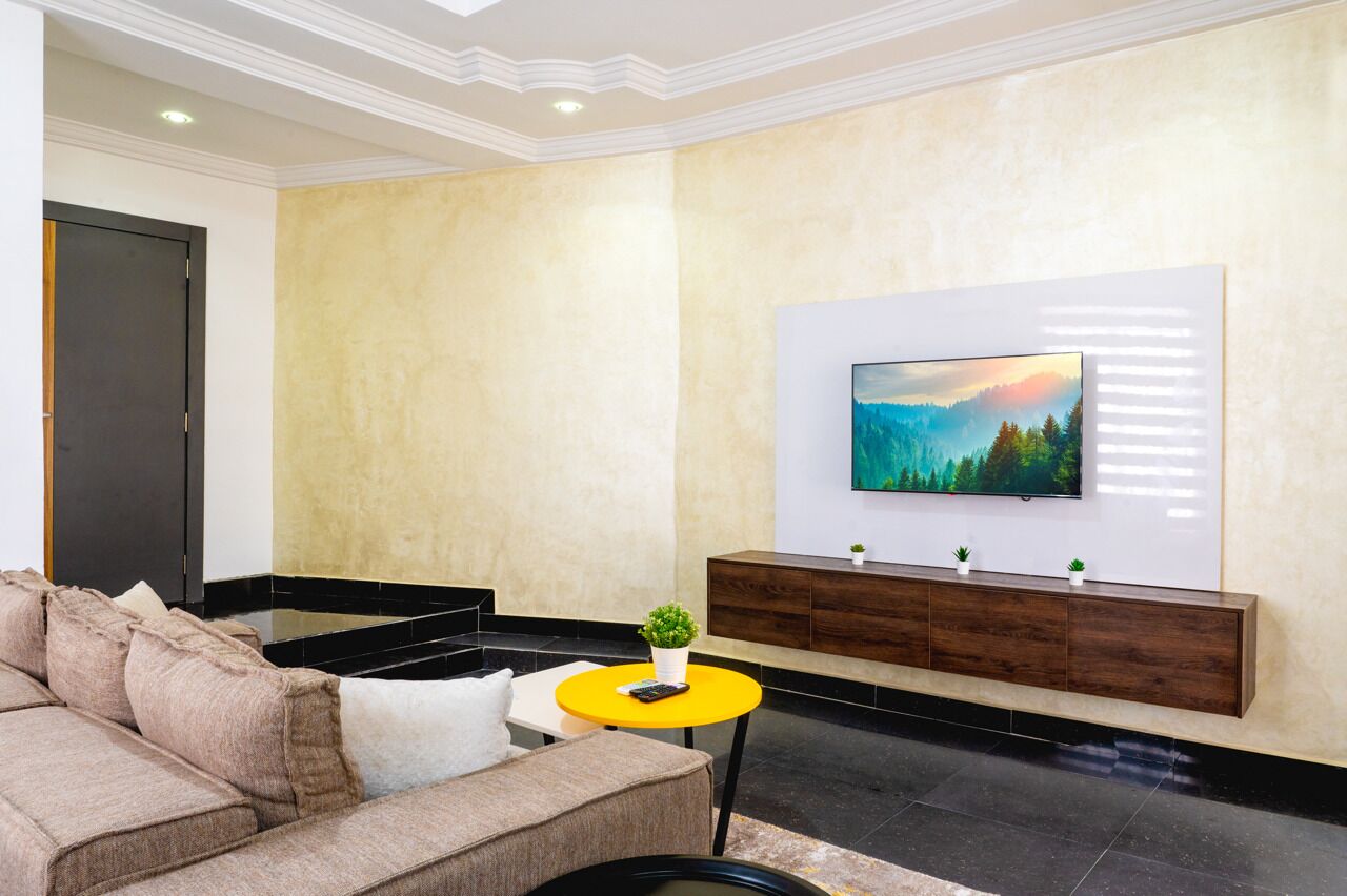 Una sala de estar moderna con pared texturizada color beige, un televisor de pantalla plana montado, un mueble bajo de madera oscura, un sofá color beige y una mesita auxiliar amarilla con una planta pequeña.