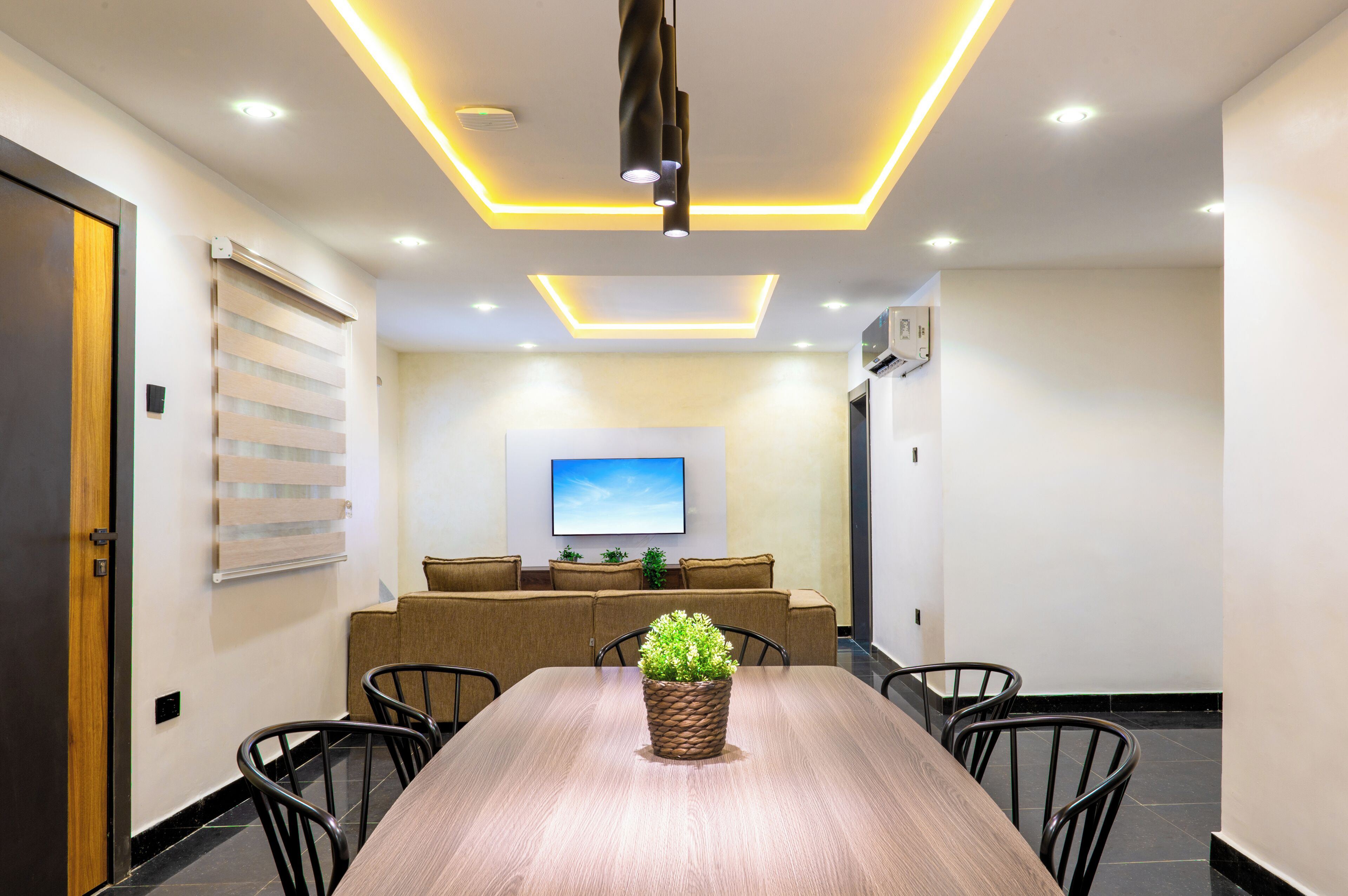 A modern dining area with a wooden table, black chairs, a potted plant, recessed ceiling lighting, and a flat-screen TV on the wall.