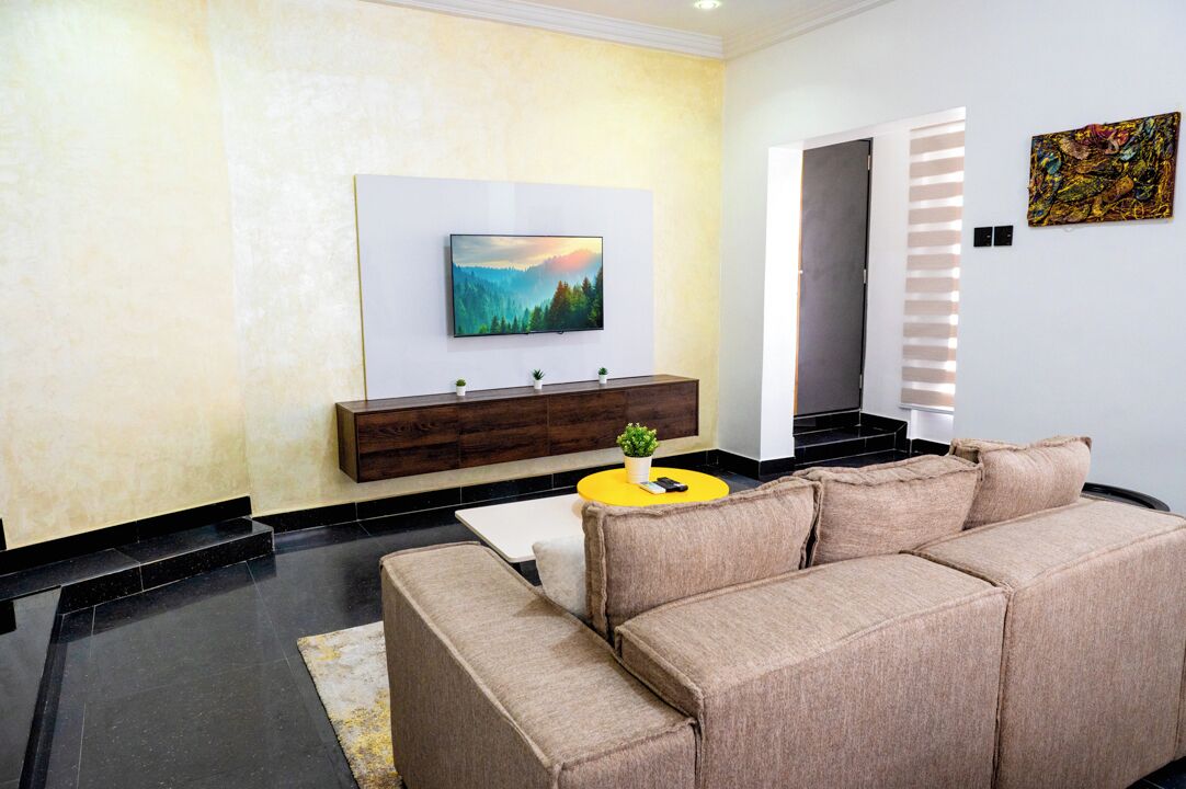 A modern living room with a beige sofa, a yellow coffee table, a flat-screen TV mounted on the wall, and a wooden console table with decorative items.