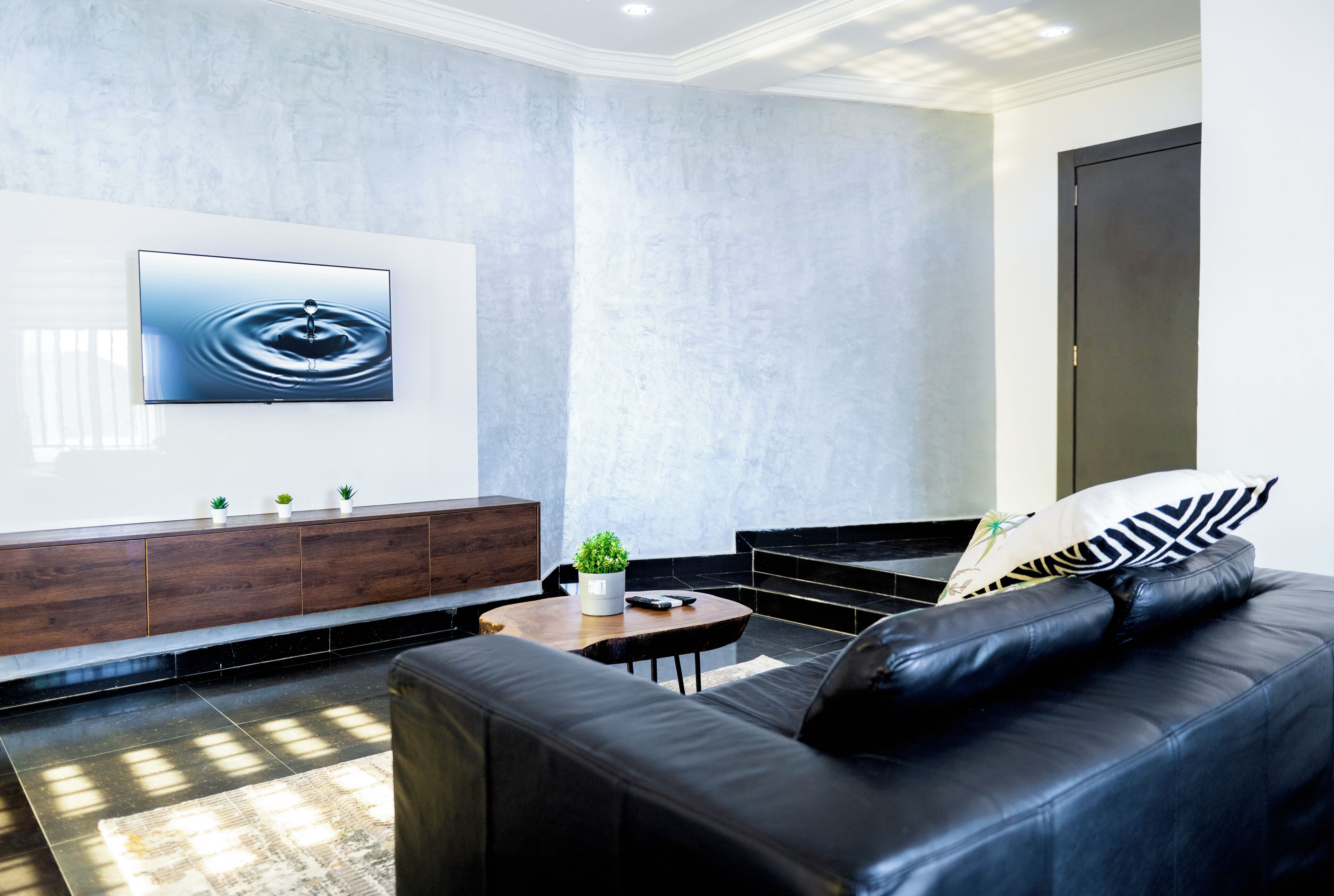 Un salon moderne avec un canapé en cuir noir, une table basse en bois, un téléviseur fixé au mur et un design minimaliste.