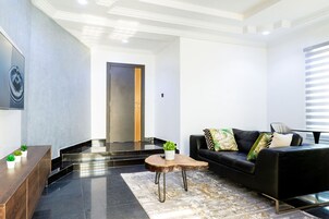 A modern living room with a black leather sofa, a wooden coffee table, and a black leather armchair. There is a large painting on the wall and a small plant on the coffee table.