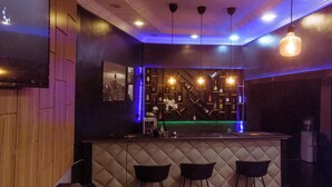 Eine Bar mit einer Theke, drei schwarzen Barhocker, einem Wand-Fernseher und einem Regal mit Flaschen und Blumen.