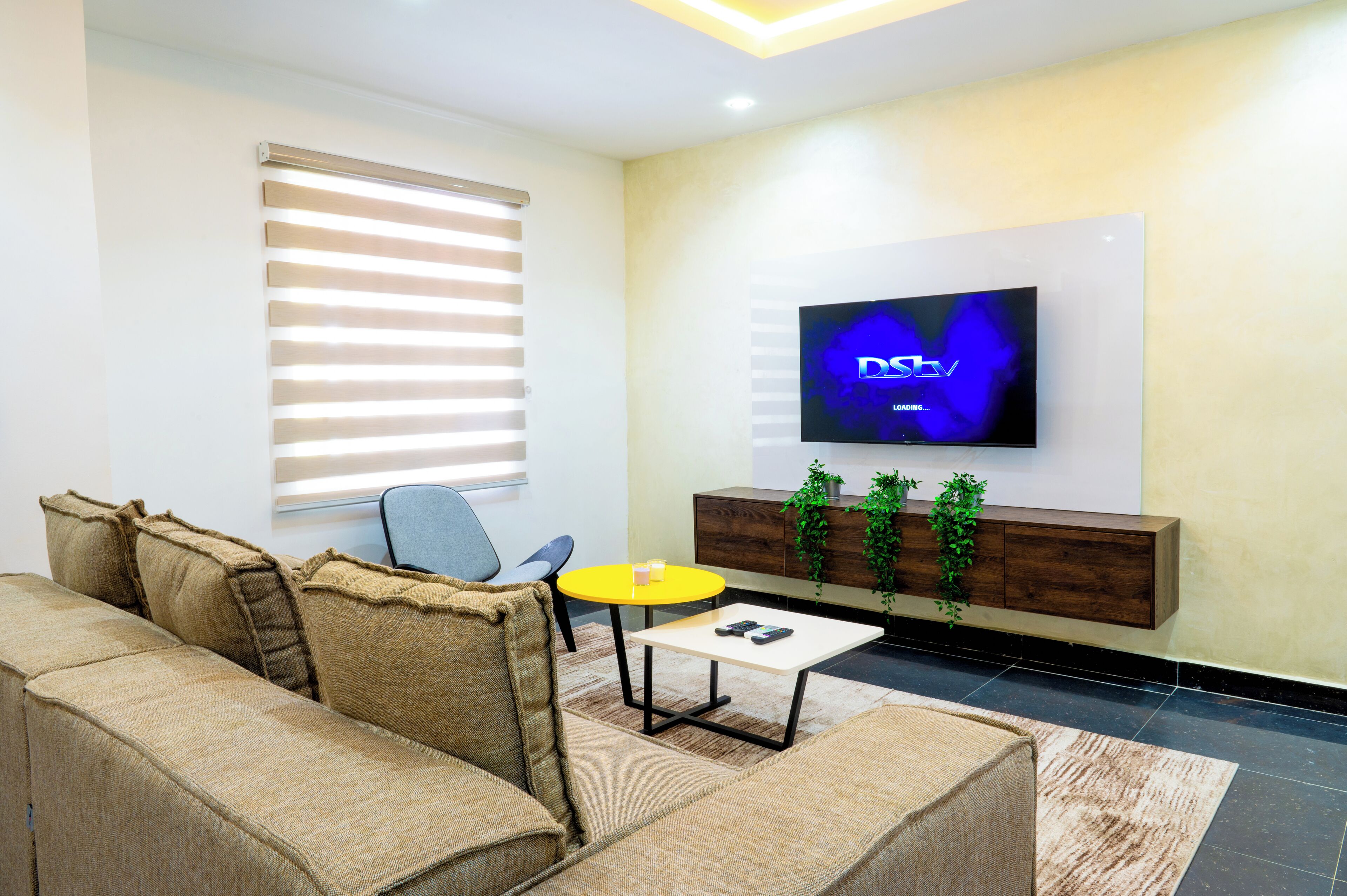 A modern living room with a sofa, a small round table, a TV mounted on the wall, and a wooden cabinet with plants.