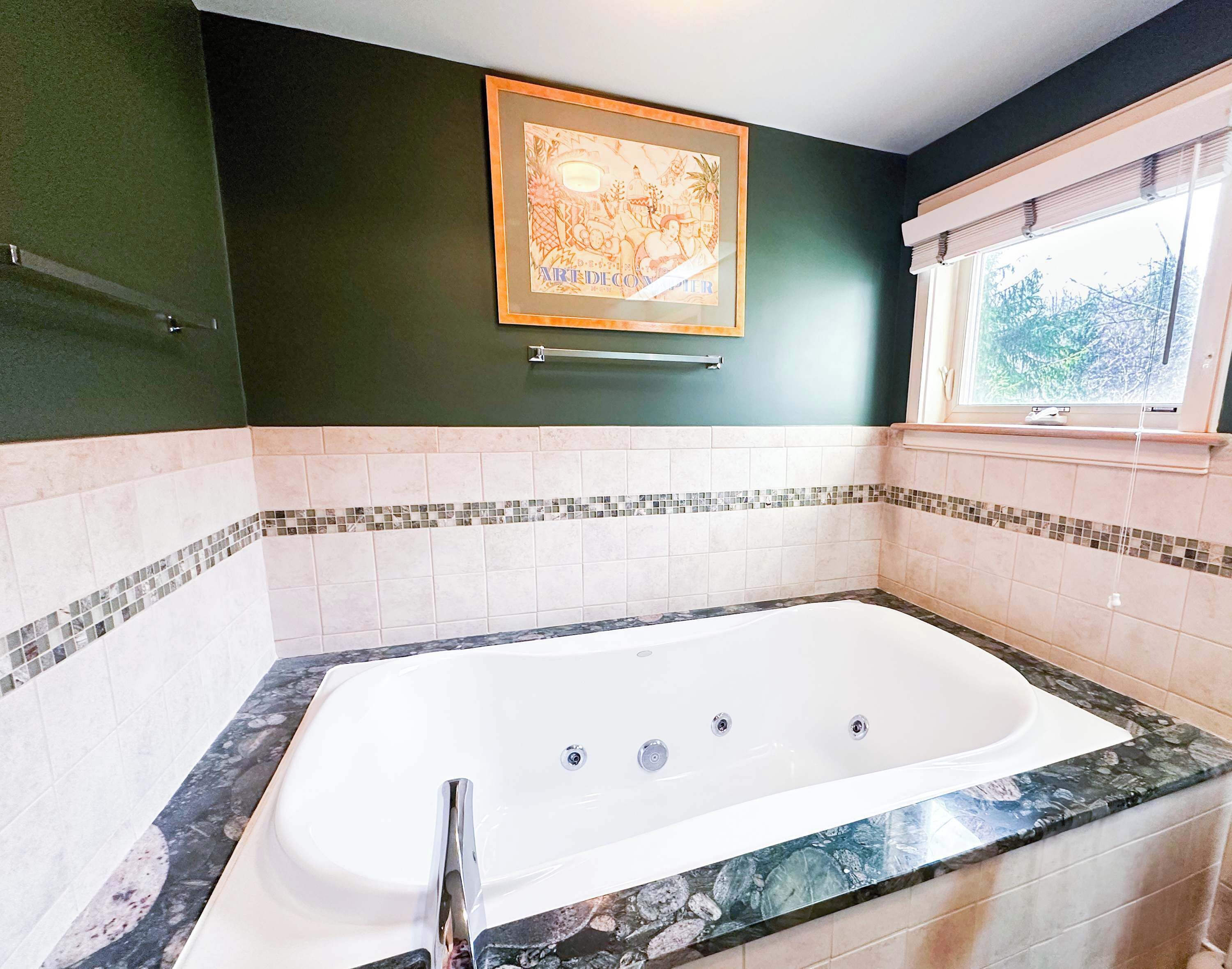 A bathroom with a large bathtub, a framed picture on the wall, and a window with blinds.