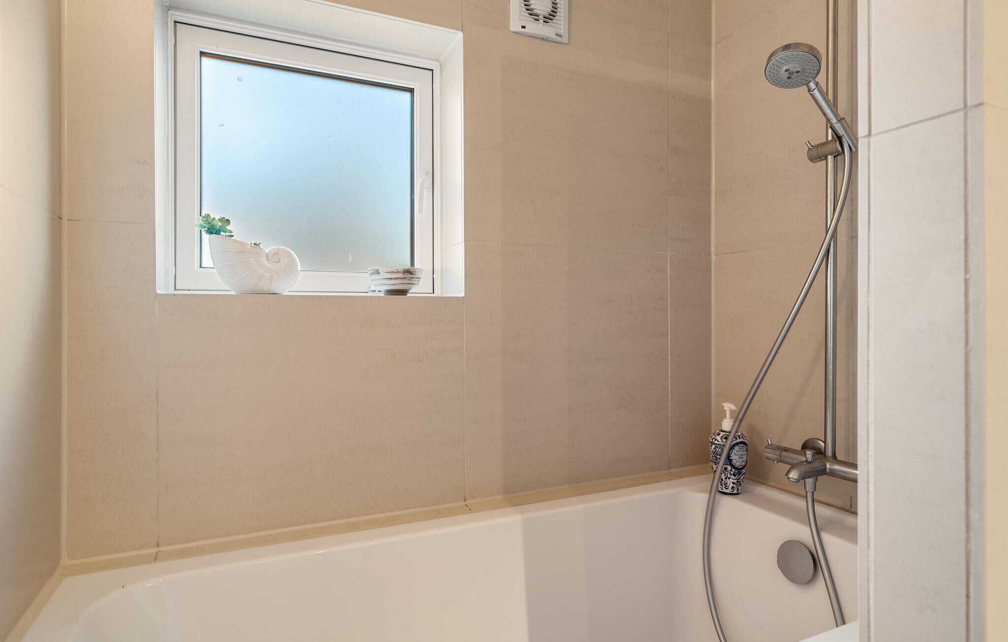 Une salle de bain avec une baignoire, une douche et une petite fenêtre agrémentée d’une plante et d’objets décoratifs.