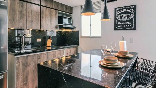 A modern kitchen with dark wood cabinets, a black countertop, and a central island. There is a microwave, a sink, and a hanging light fixture above the island.