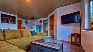 A cozy living room with a sofa, a coffee table, and a TV. There's a kitchen in the background with cabinets and a fridge.