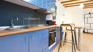 Une cuisine moderne avec des armoires bleues, un plan de travail en bois, un évier en acier inoxydable, un four à micro-ondes et une cuisinière. On y trouve une chaise et une table, ainsi qu’un luminaire suspendu.