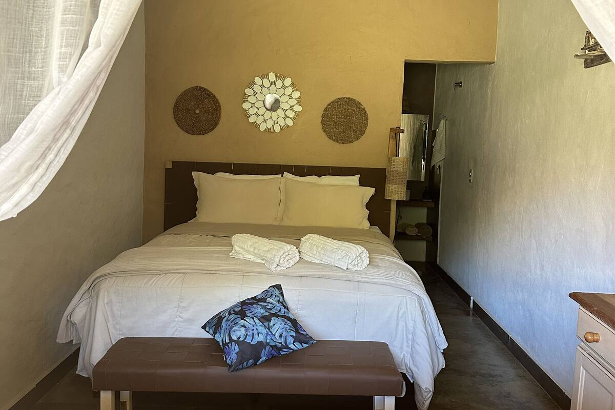 A bedroom with a bed, a bench, a wall-mounted mirror, and a door leading to another room.