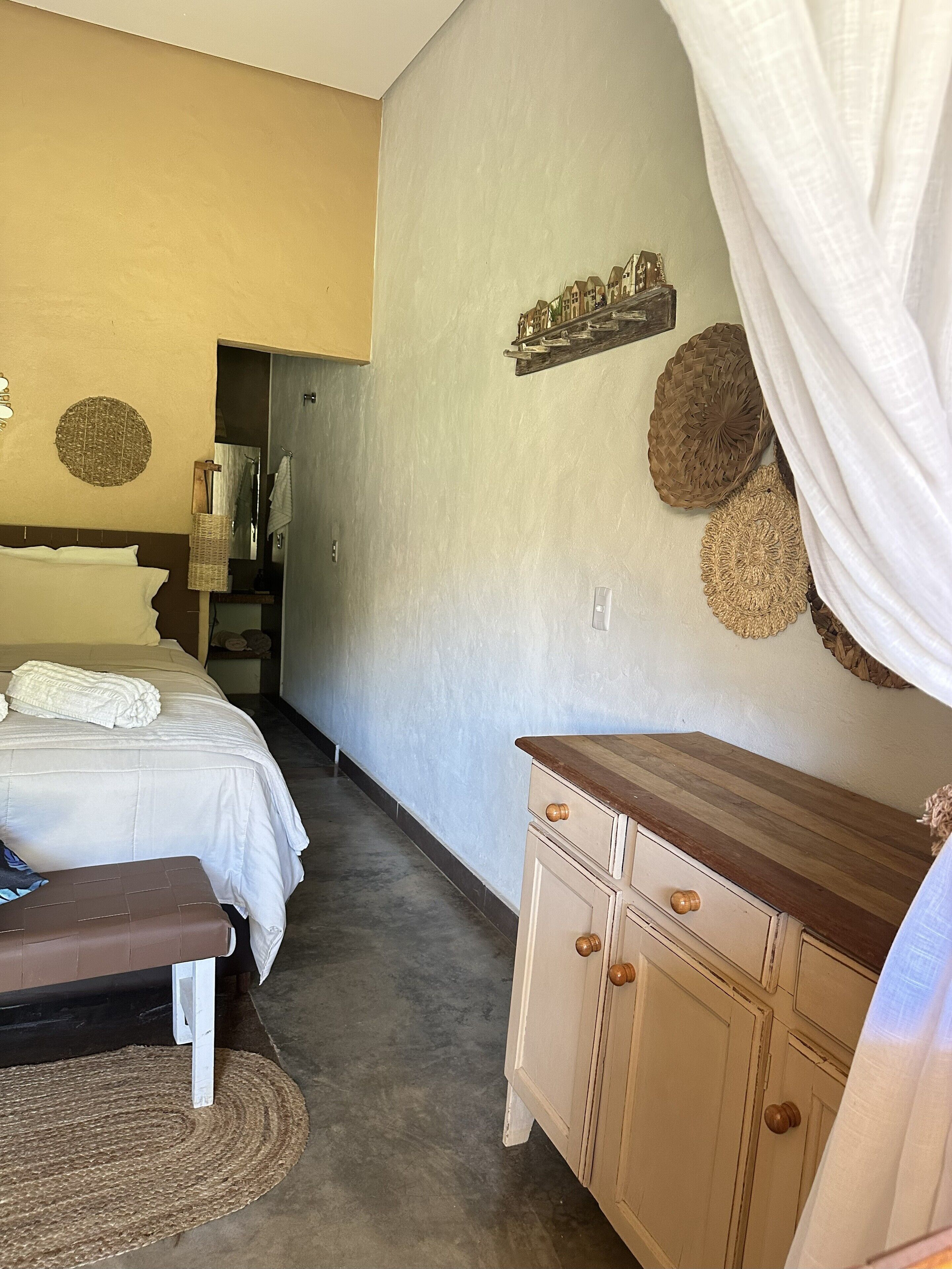 Un dormitorio con cama, mesita de noche y cómoda. Hay un espejo, una toalla y una repisa con objetos decorativos.