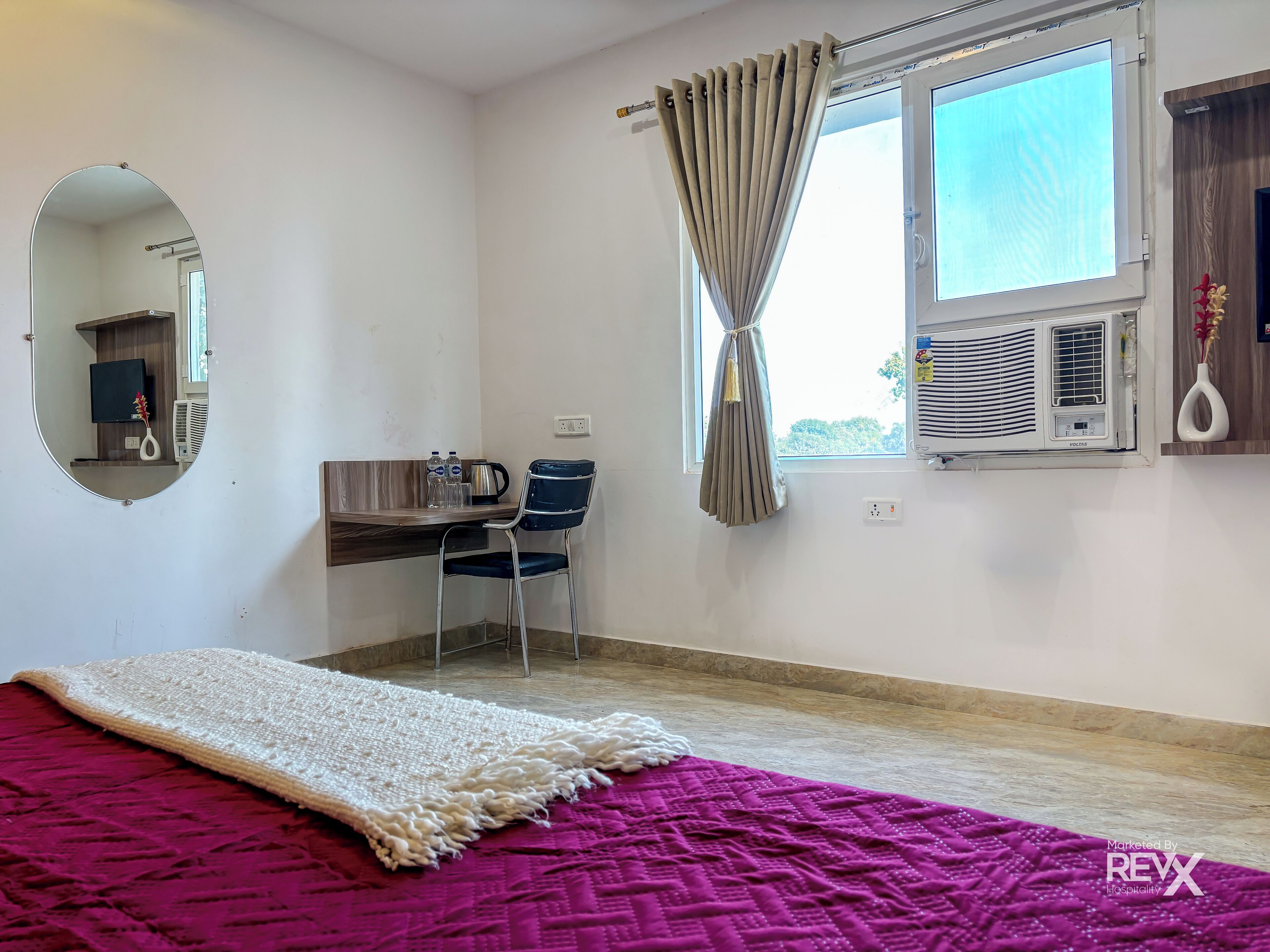 A bedroom with a bed, a desk, a chair, a mirror, a window with curtains, an air conditioner, and a wall-mounted shelf.